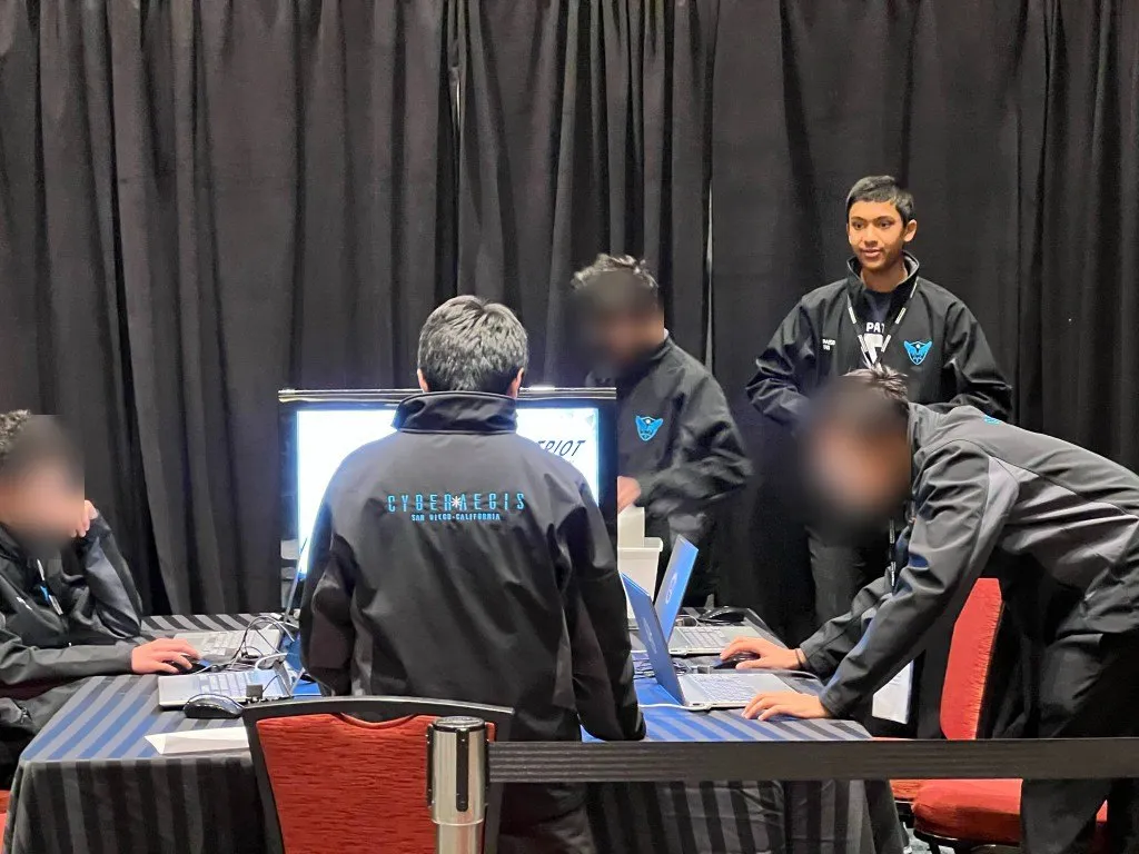 Aditya standing with CyberAegis at the finals table during the live round; teammates' faces blurred