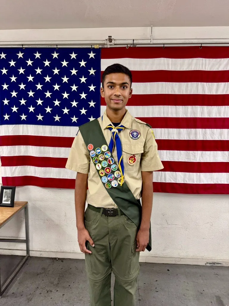 Aditya in his Boy Scout uniform and merit badge sash in front of an American flag