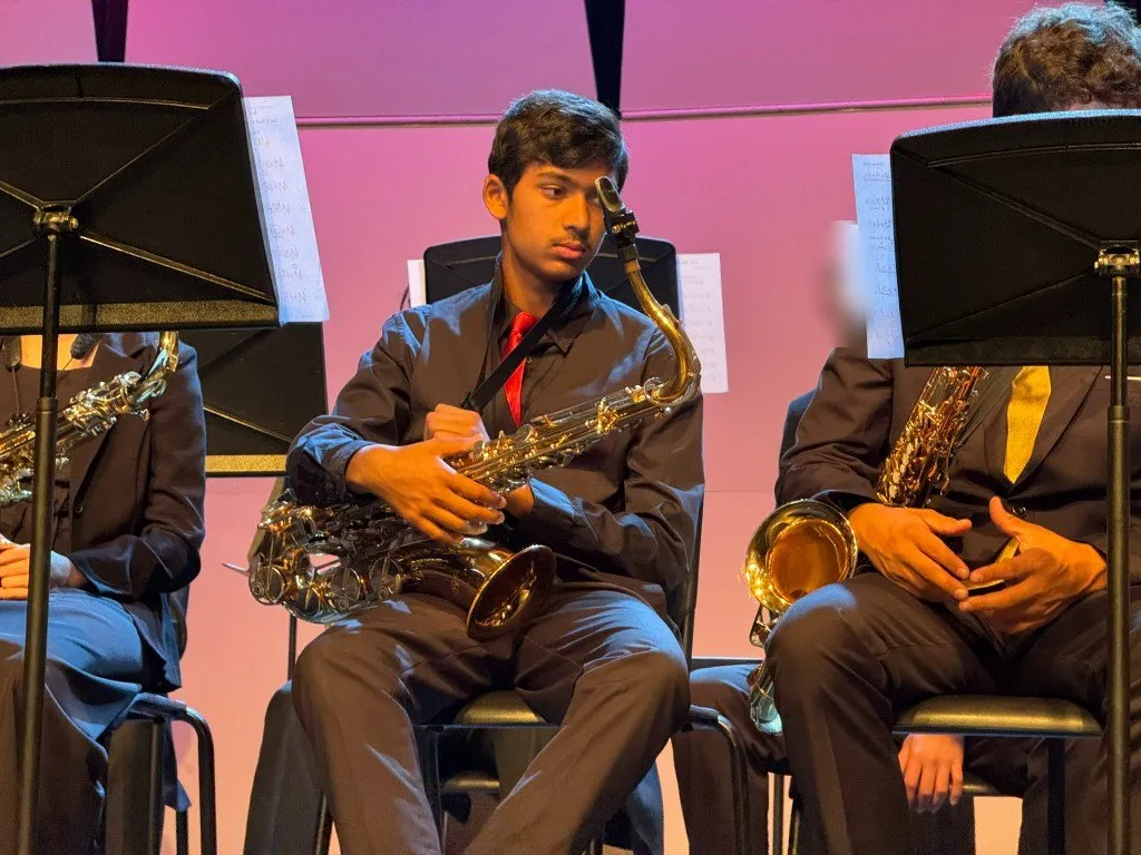 Aditya seated on stage with his tenor saxophone in a red tie