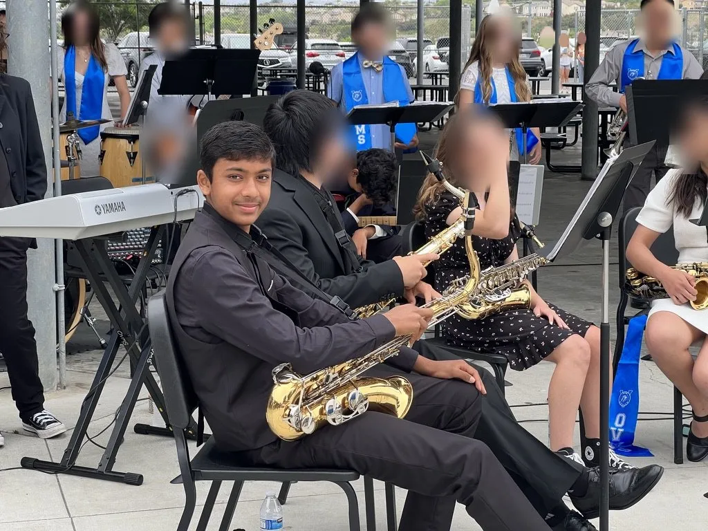 Aditya smiling with his tenor saxophone at an outdoor concert; other faces blurred